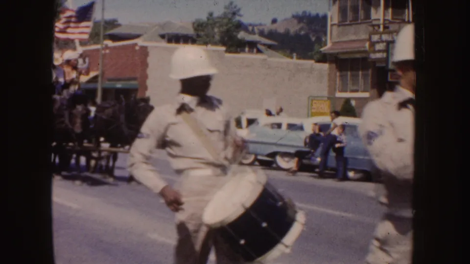 1954 South Dakota parade frame with a drummer in a white helmet, an American flag, spectators, storefronts, and mid-century cars in the background.