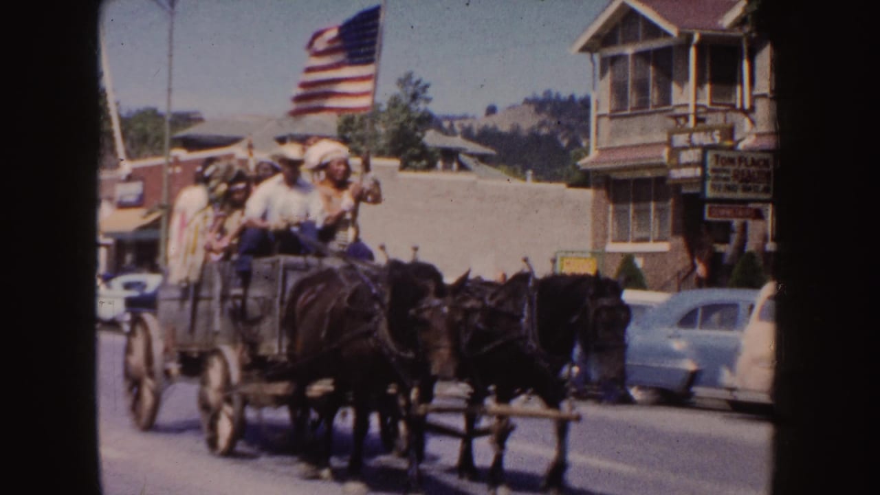 1954 South Dakota home-movie frame showing a horse-drawn wagon with riders, an American flag, parked cars, and town storefronts.