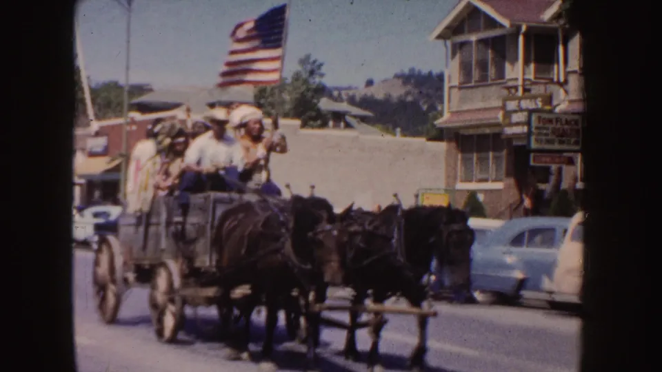 1954 South Dakota home-movie frame showing a horse-drawn wagon with riders, an American flag, parked cars, and town storefronts.
