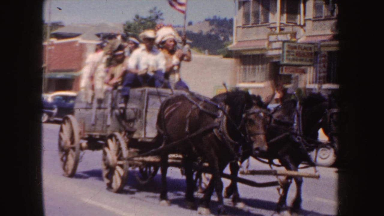 1954 South Dakota parade frame showing two horses pulling a wagon of riders past storefronts, parked cars, and street-side signage.