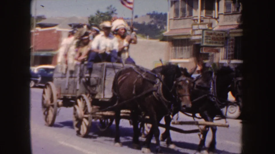 1954 South Dakota parade frame showing two horses pulling a wagon of riders past storefronts, parked cars, and street-side signage.