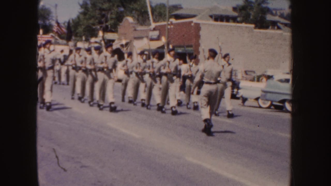 Uniformed marchers carrying rifles move down a sunlit street past parked cars and brick buildings.