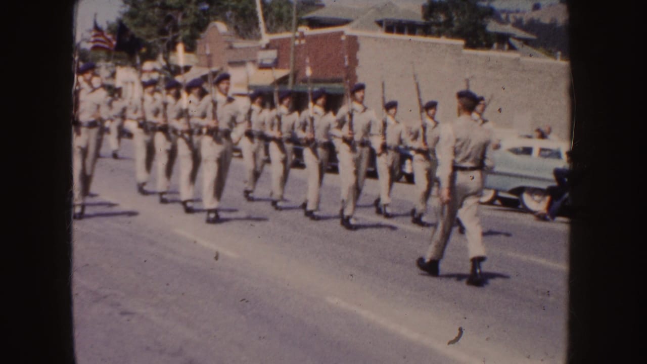 Uniformed marchers in a 1954 South Dakota parade carry rifles while a leader walks beside the formation near parked mid-century cars.