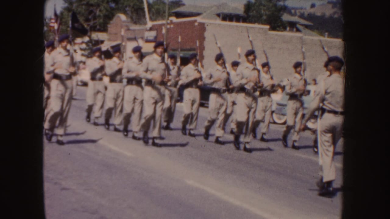 The 8mm frame shows tan-uniformed marchers with rifles moving close to the roadside camera during a South Dakota parade scene.