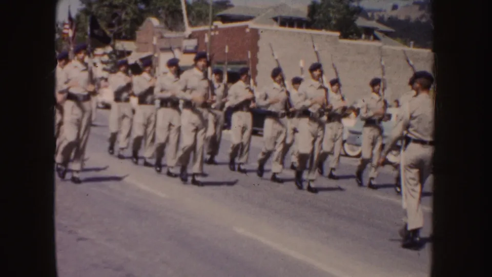The 8mm frame shows tan-uniformed marchers with rifles moving close to the roadside camera during a South Dakota parade scene.