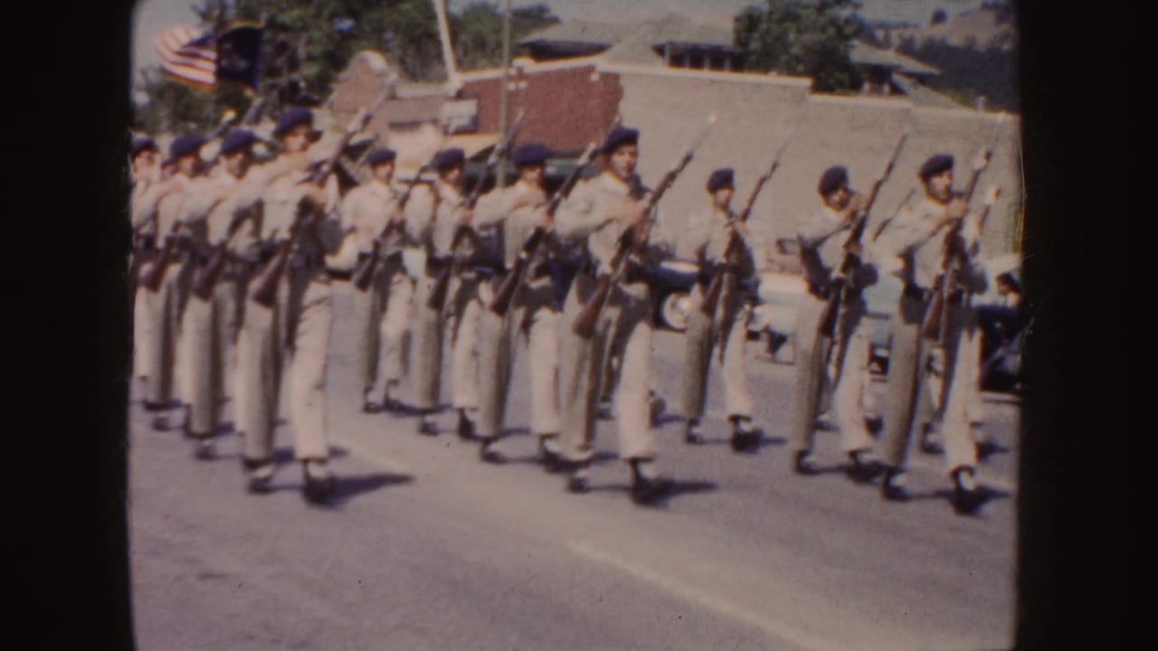 Near the end of the clip, tan-uniformed marchers carry upright rifles as an American flag and parked cars appear behind them.