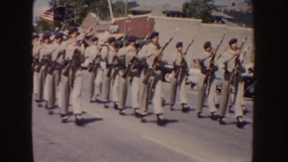 Near the end of the clip, tan-uniformed marchers carry upright rifles as an American flag and parked cars appear behind them.