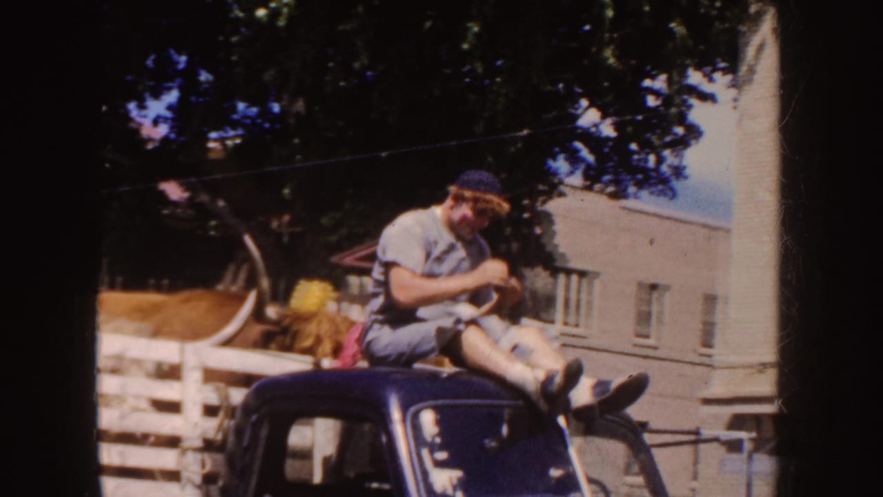 Young man in a blue cap sits on the roof of a dark vintage car beside a livestock trailer.