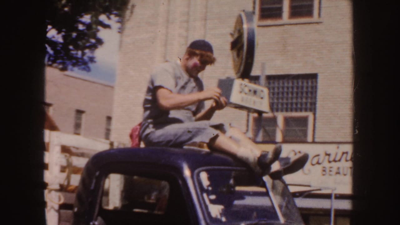A young parade participant in casual clothes sits on a dark car roof as a horned steer in a trailer passes through a South Dakota street scene.