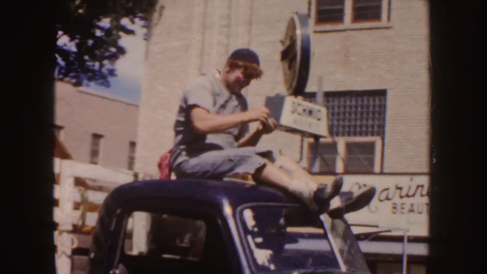 A young parade participant in casual clothes sits on a dark car roof as a horned steer in a trailer passes through a South Dakota street scene.