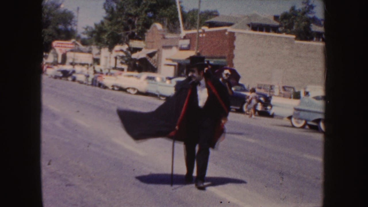 A man dressed in a black suit, top hat, and cape walks with a cane during the 1954 South Dakota parade, with vintage cars behind him.