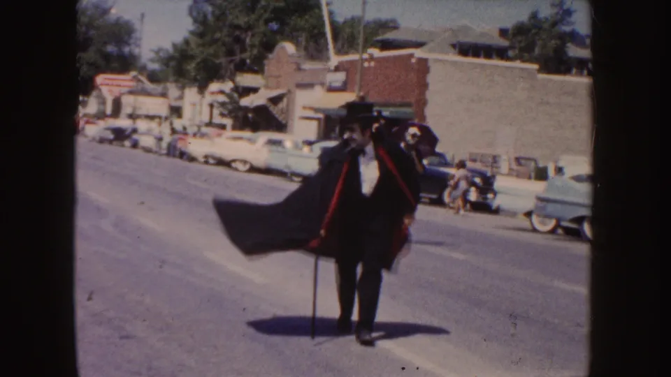 A man dressed in a black suit, top hat, and cape walks with a cane during the 1954 South Dakota parade, with vintage cars behind him.