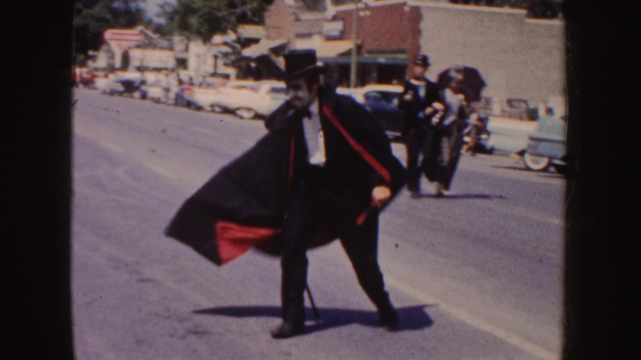The caped man in top hat and cane moves across a South Dakota parade route, with other costumed figures and 1950s cars visible behind him.