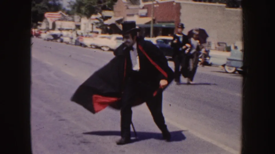 The caped man in top hat and cane moves across a South Dakota parade route, with other costumed figures and 1950s cars visible behind him.