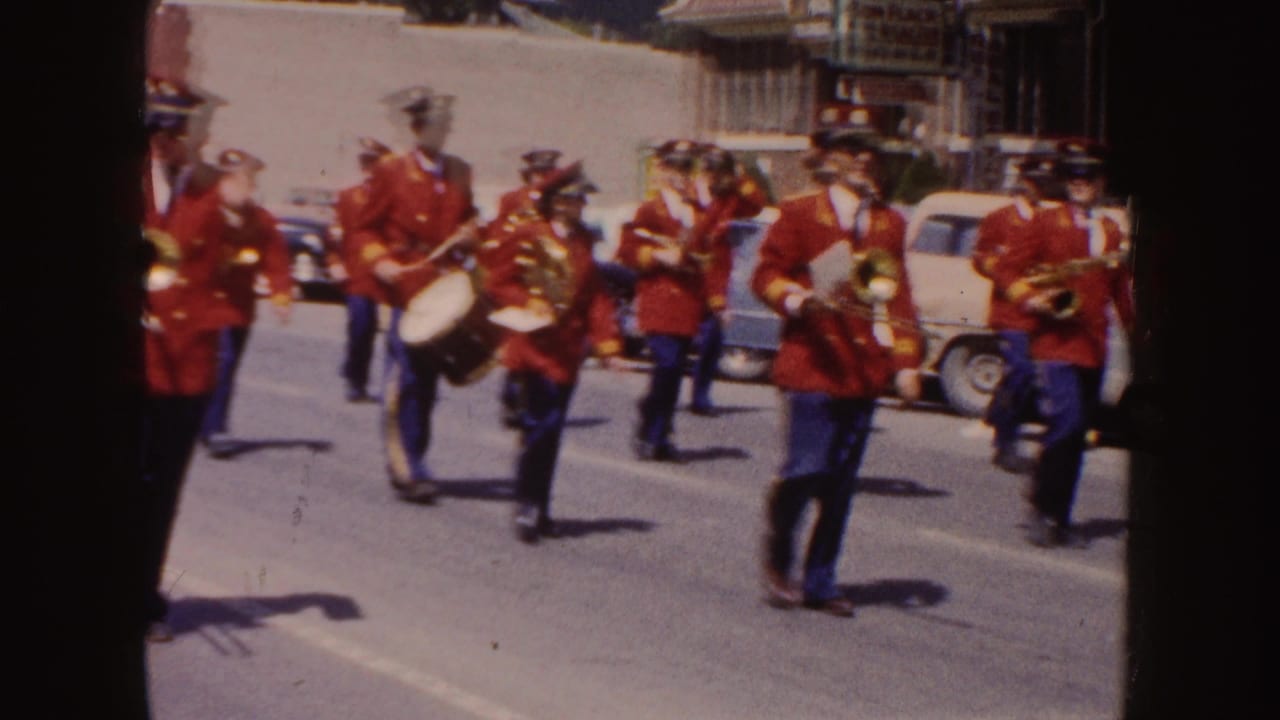 Marching band in red coats and blue pants advances down a sunlit street.