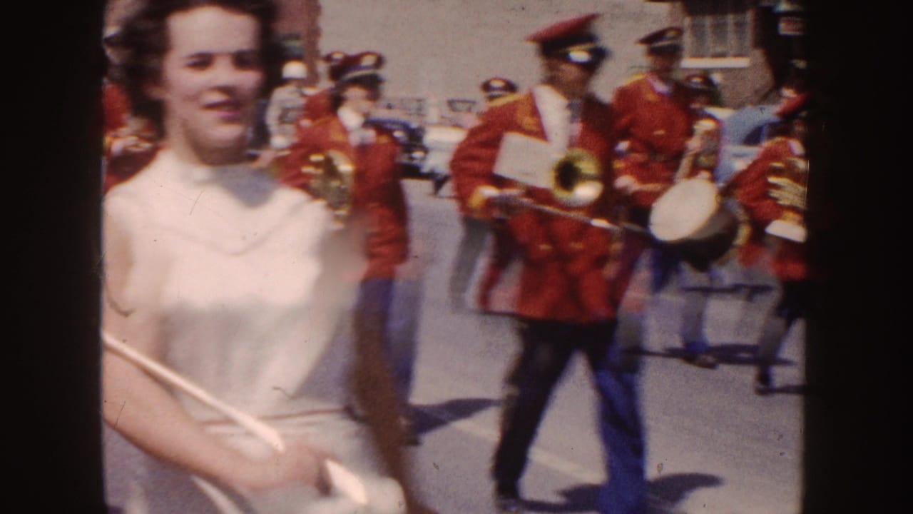 A woman in a white dress passes close to the camera as band members in red coats continue marching behind her.