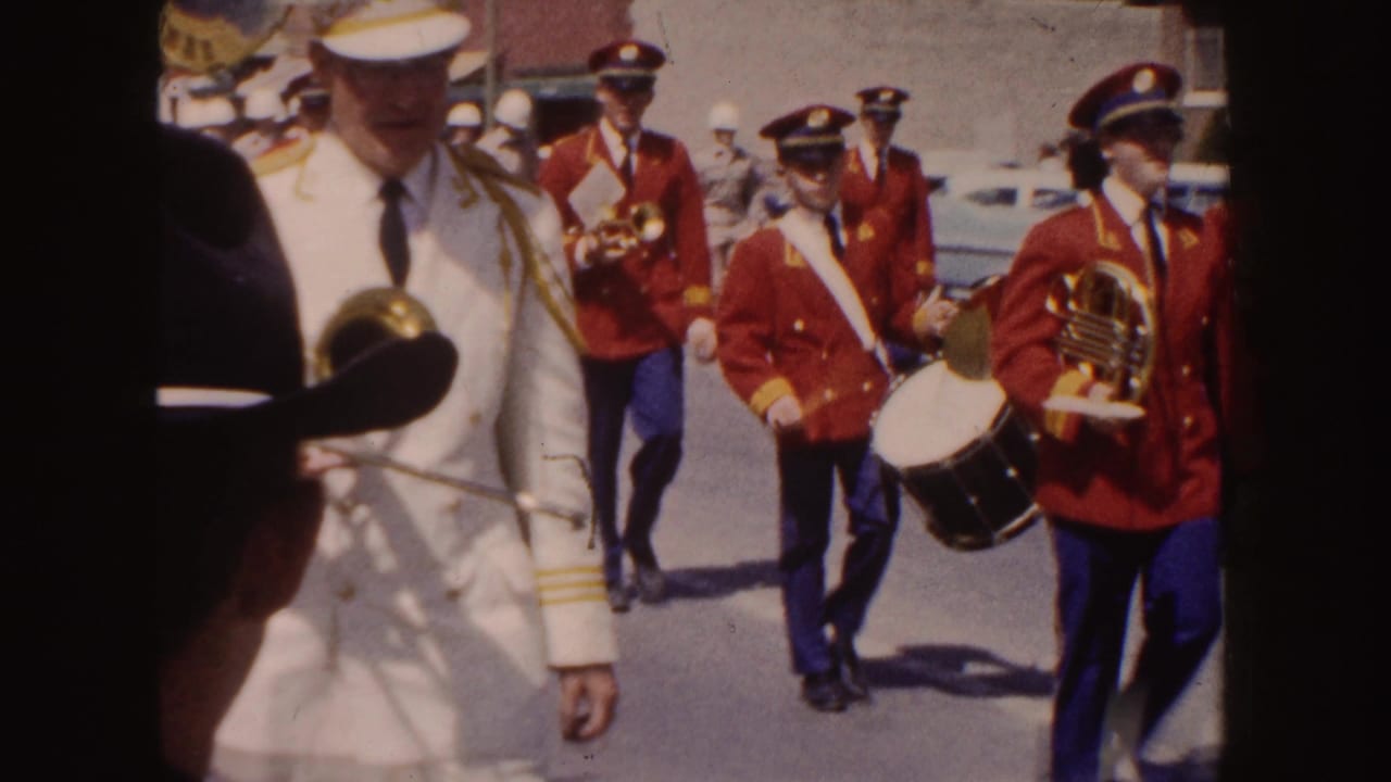 Parade participants include a man in a white uniform near the camera and red-coated musicians with drum and horn behind him.