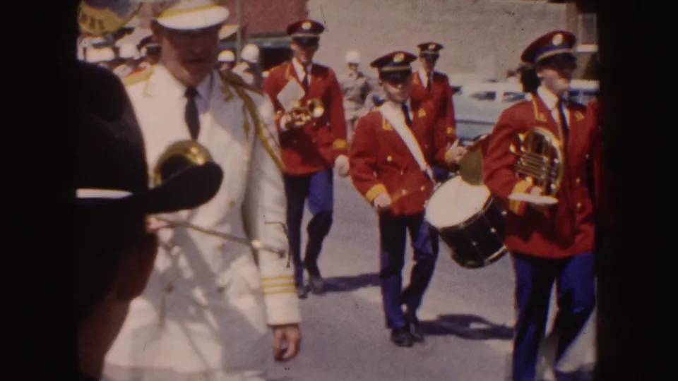 Parade participants include a man in a white uniform near the camera and red-coated musicians with drum and horn behind him.