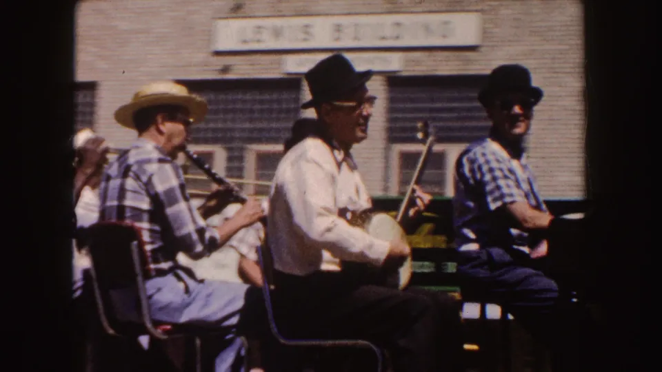 Outdoor folk musicians sit near a brick building, including a banjo player and a clarinet player, with signage above them.
