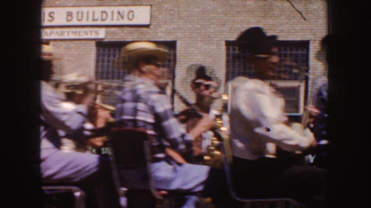1954 South Dakota 8mm frame of brass band musicians seated on a city street near an apartment building sign.