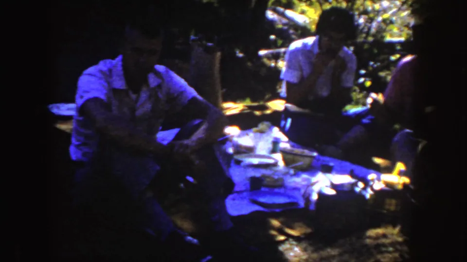 1954 South Dakota 8mm frame of three people eating around a shaded picnic table in a wooded outdoor area.