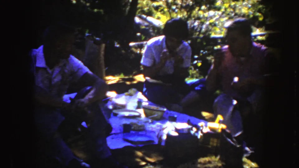 1954 South Dakota home-movie frame of a small shaded picnic meal with three seated people and table items visible.