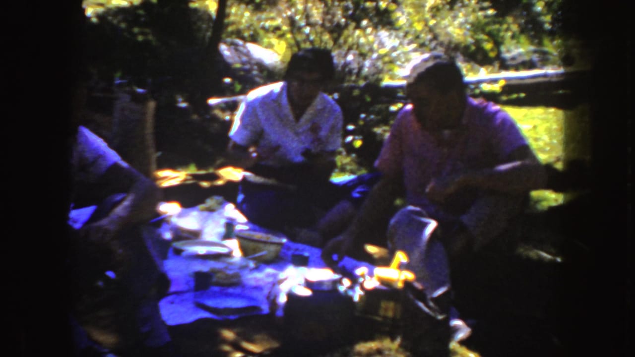 1954 South Dakota home-movie frame showing adults seated around a picnic blanket under trees, with food and containers in view.