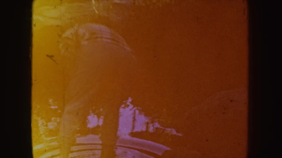 In South Dakota 1954 home-movie footage, a woman bends near shallow water, partly obscured by heavy orange color shift.