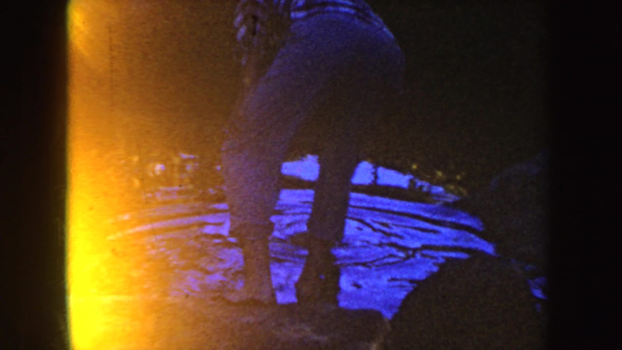 The woman remains at the stream edge in the 1954 South Dakota footage, seen through yellow-blue color drift and dark frame edges.