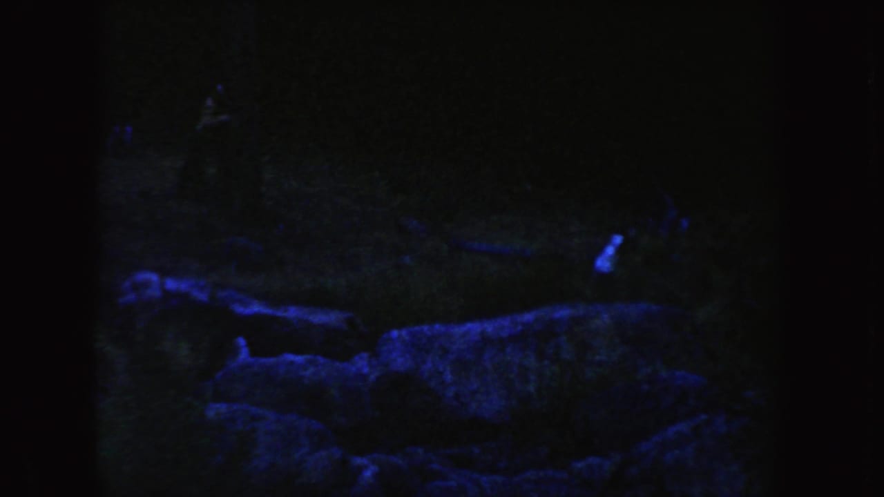 1954 South Dakota 8mm frame showing a dim blue-lit rocky or earthen ledge with indistinct people at a distance.