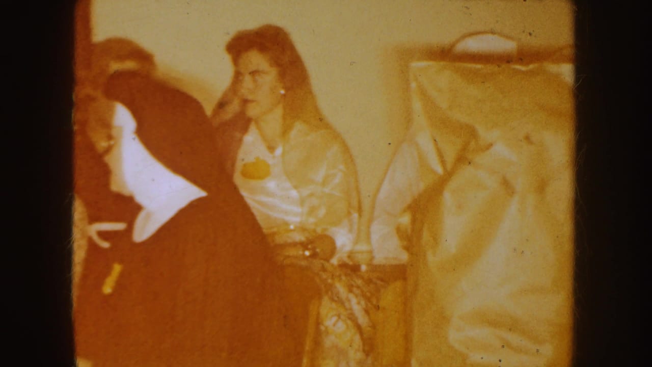 South Dakota 1954 home-movie frame of women seated indoors, with one woman in a light blouse facing left beside a large coat or hat.