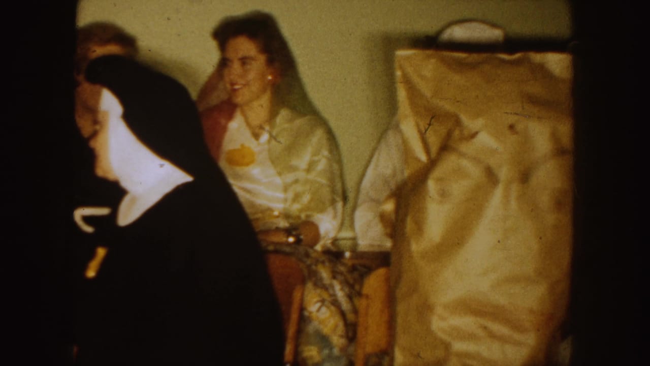 1954 South Dakota 8mm frame showing a nun in a black habit near women and paper-covered seated figures during an indoor religious gathering.