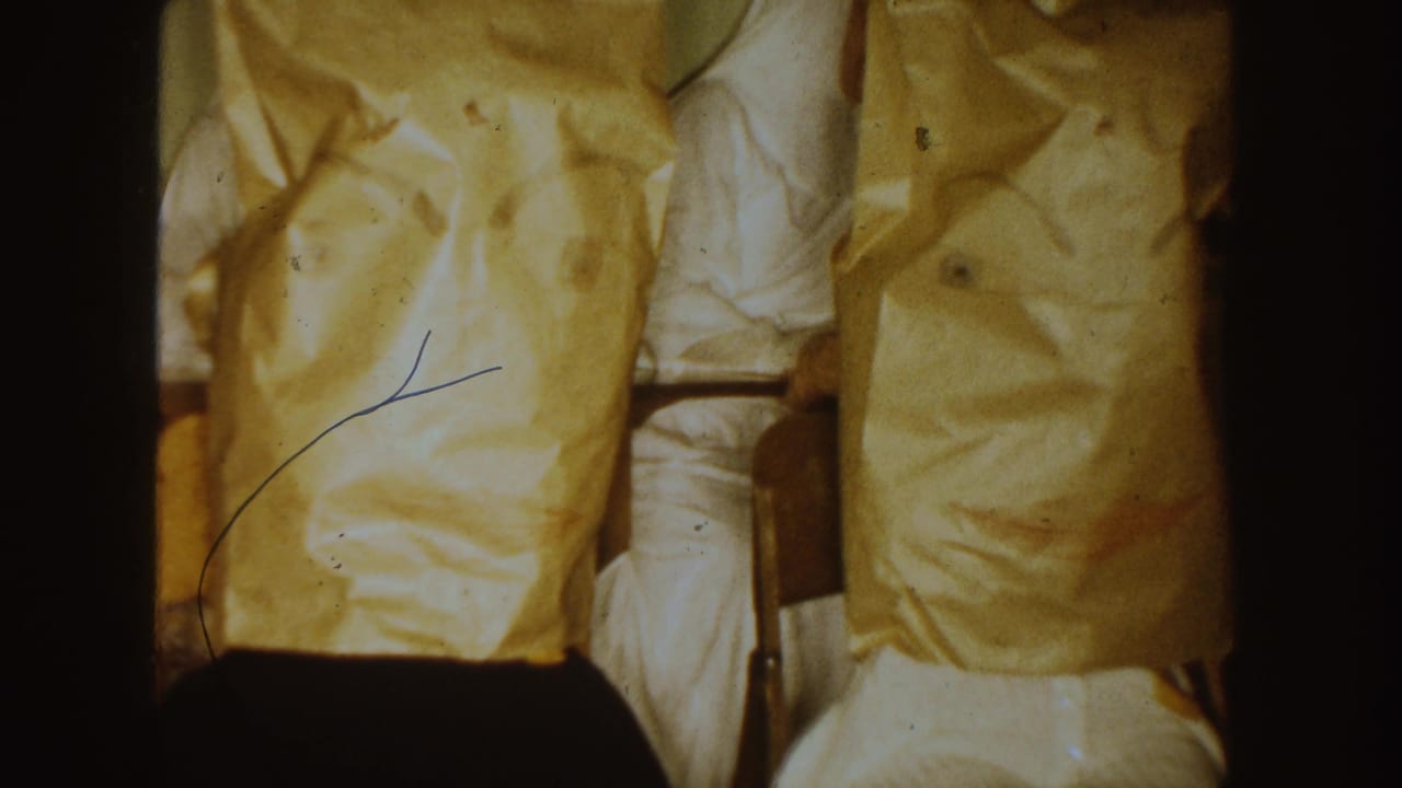 1954 South Dakota 8mm frame with two paper-covered seated figures and a partly visible person in white clothing inside a religious setting.