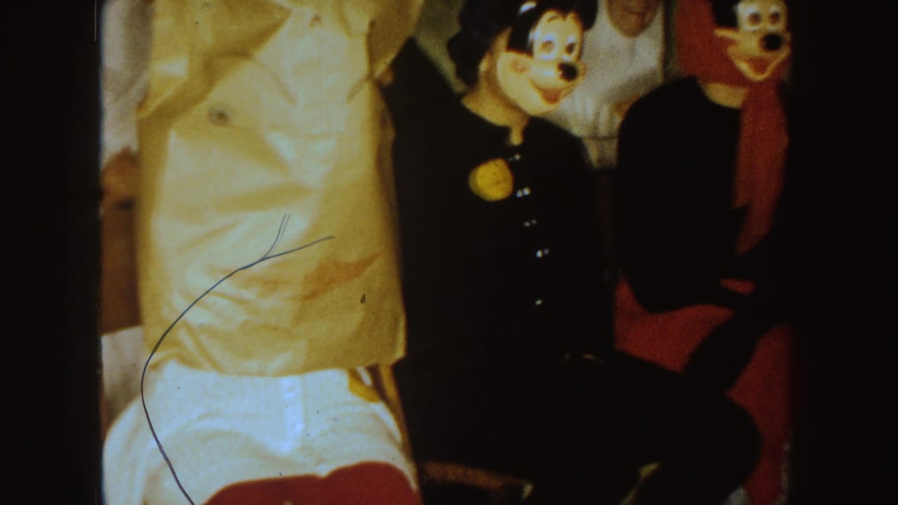 Masked party guests seated indoors, including a Mickey-style character mask beside a figure in a red headscarf.