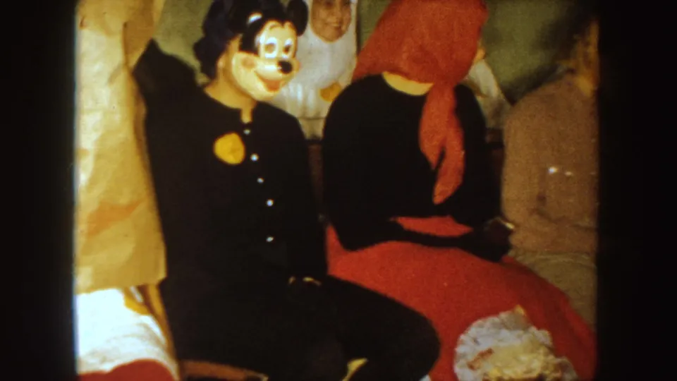 1954 South Dakota 8mm frame showing a Mickey-style masked adult, a seated figure in a red headscarf, and other costumed guests gathered indoors.