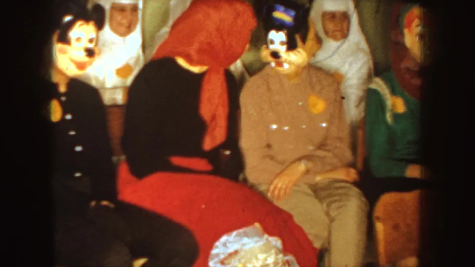 1954 South Dakota home-movie frame showing seated party guests facing the camera, with Mickey- and Goofy-style character masks, a red headscarf, and a green garment at the edge.