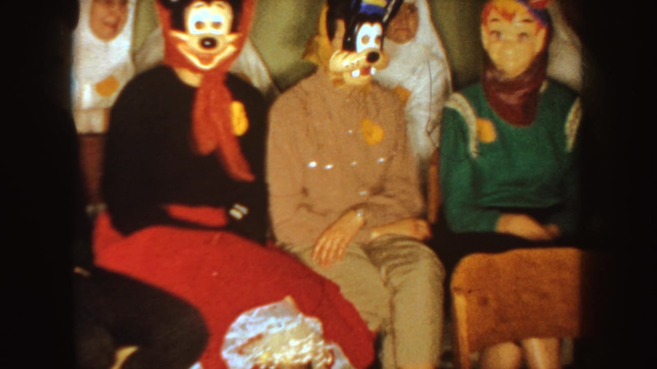 1954 South Dakota home-movie frame of seated adults at an indoor Halloween party with character masks and costumes.
