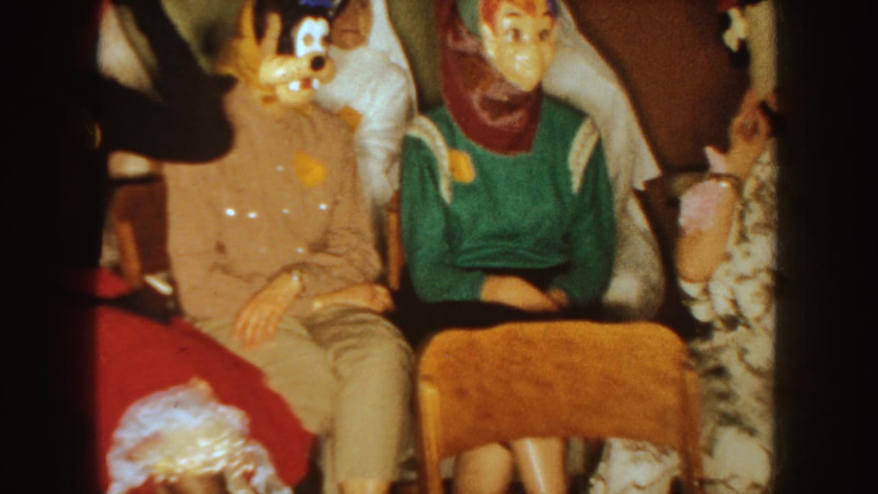 Seated adults in masks and costumes, with a woman in a green sweater near the center.