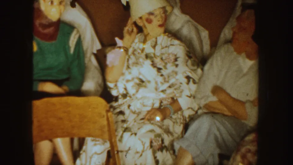 1954 South Dakota frame of adults seated closely at an indoor Halloween party with costumes and masks visible.