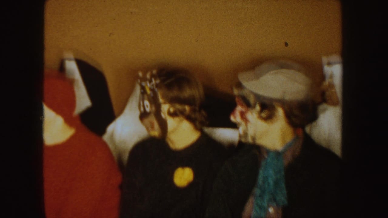 Seated costumed adults, including a red hood, black animal mask, and painted face, in a dim room.