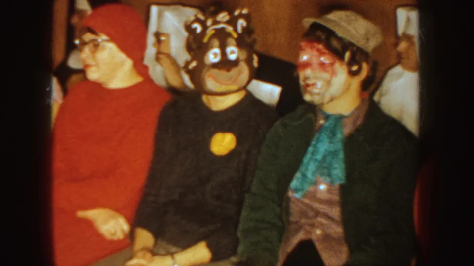 1954 South Dakota home-movie frame of seated Halloween guests in red clothing, an animal mask, and theatrical face paint.