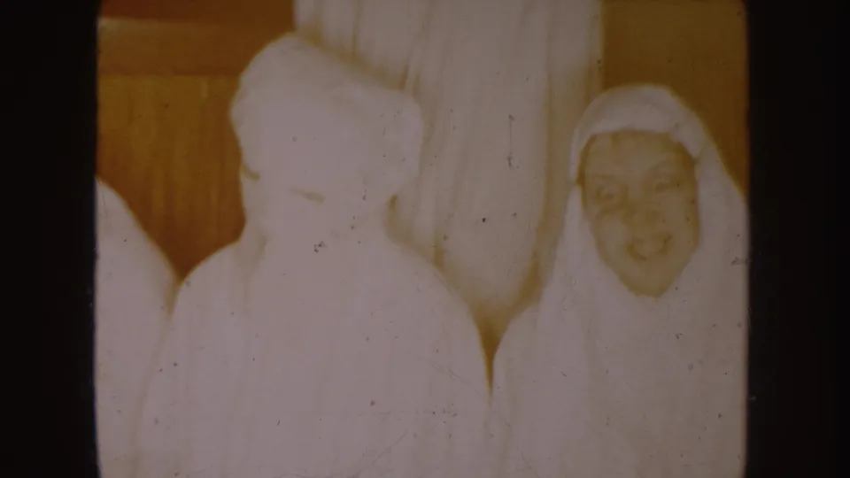 1954 South Dakota 8mm frame showing white-clad figures with veils or robes near a taller robed figure inside a private interior.
