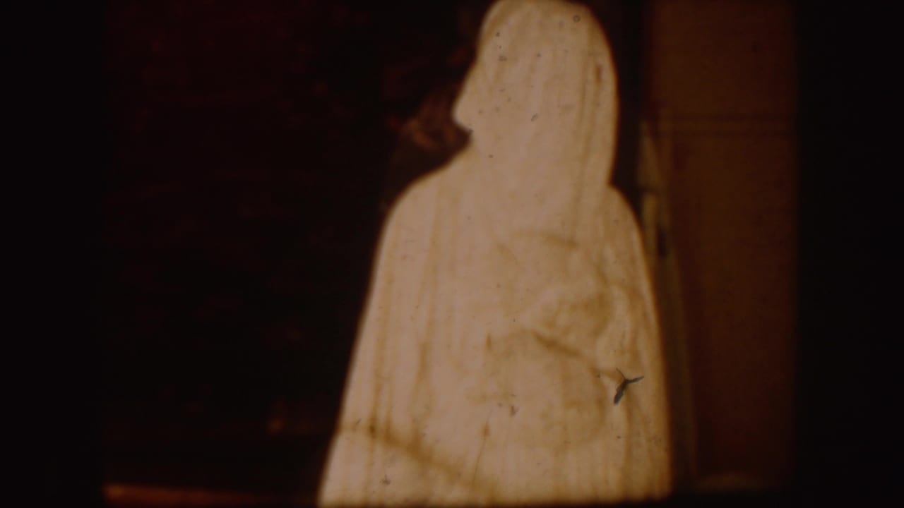 Tall white robed figure standing against a dark interior background.