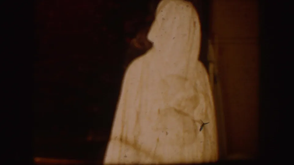 Tall white-robed figure appears in a dim South Dakota home-movie interior, with soft focus and warm aged film color.