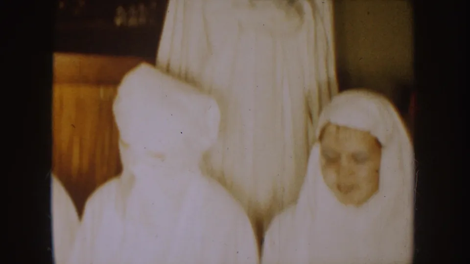 1954 South Dakota home-movie frame showing a taller robed figure behind shorter white-clad figures, including a visible face, indoors.