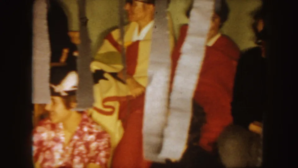 1954 South Dakota 8mm frame showing a costumed partygoer and seated adults in a decorated indoor gathering.