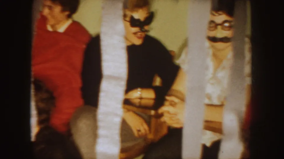 1954 South Dakota home-movie frame with two adults in novelty masks seated beside another guest at an indoor party.