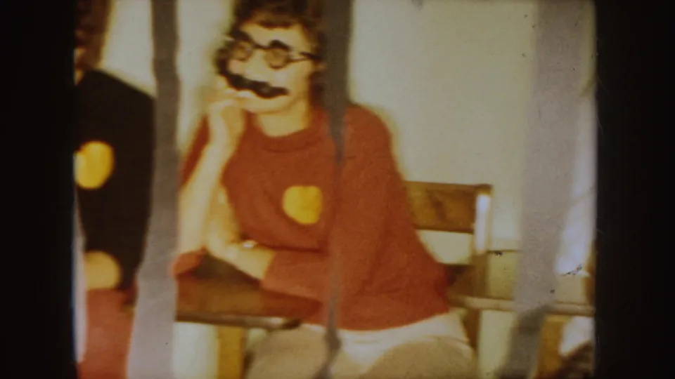 Student in a red shirt sits at a classroom desk with fake mustache and glasses in the South Dakota school-party home movie.
