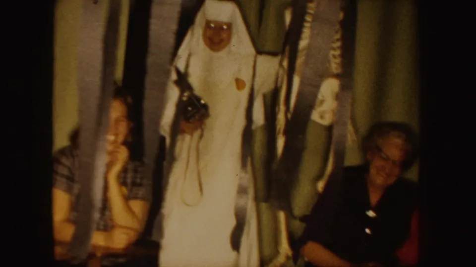 1954 South Dakota home-movie frame showing a white-costumed adult near seated adults in a private indoor room.