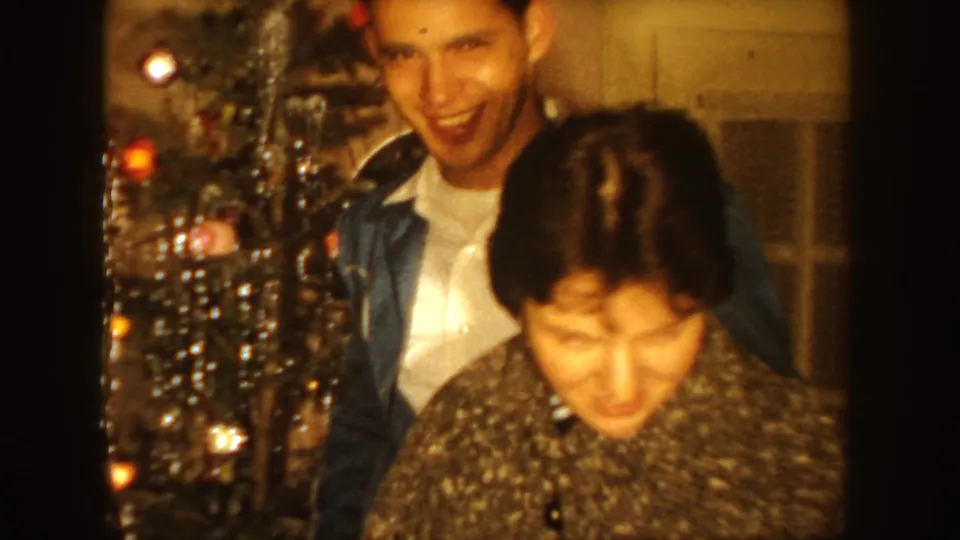 Young couple in a 1954 South Dakota home interior, smiling and standing near a decorated Christmas tree.